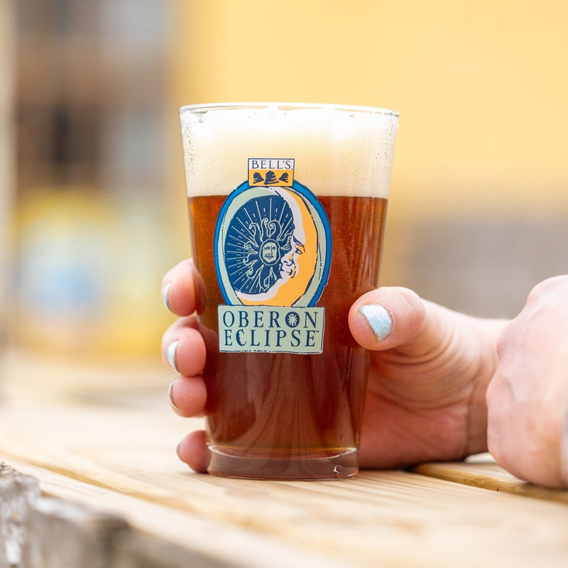 A clear glass 16oz pint with a Bell's Oberon Eclipse logo, featuring a yellow crescent moon with a face and an Oberon Sun. 