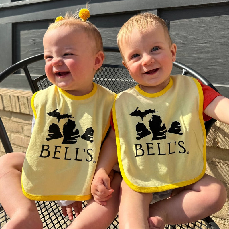 Light yellow baby bib with bold yellow lining and modified Bell's logo in black.  The middle Bell in the logo is replaced by the silhouette of the state of Michigan.