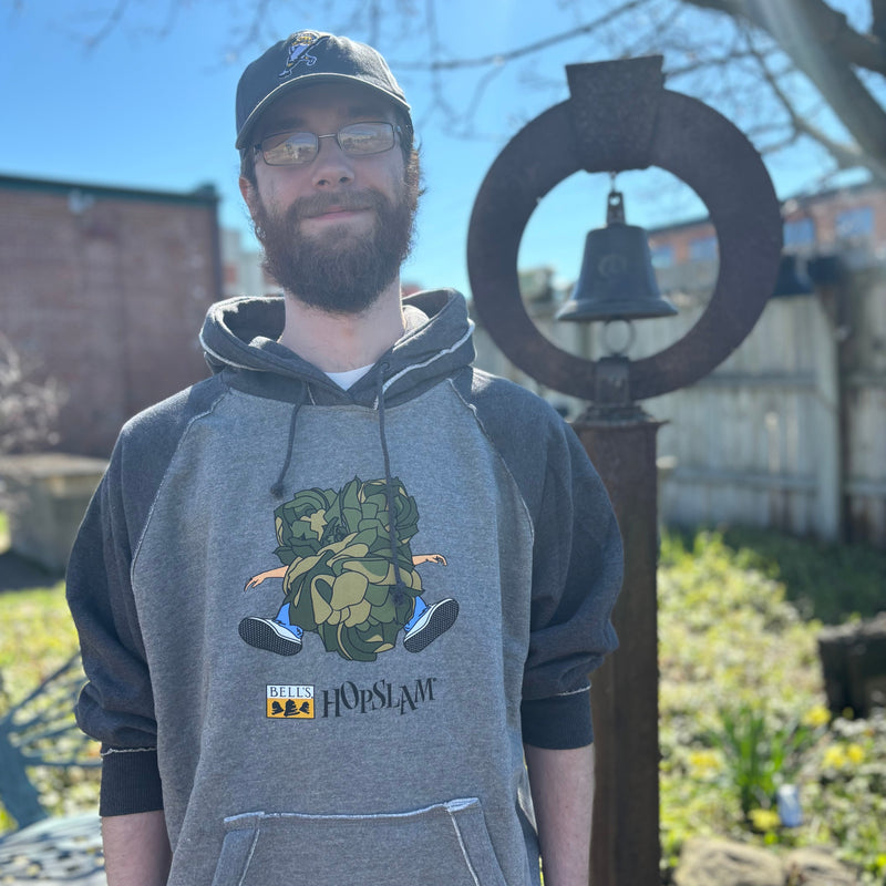 Gray hoodie with black sleeves, hood, and drawstrings. Printed on the front is the Bell's Brewery Hopslam logo underneath an illustration of 3 large hop plants slamming into a person in jeans and sneakers.