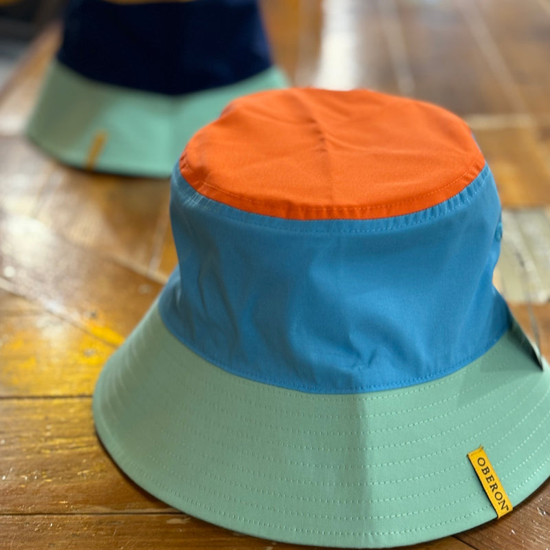 A reversible bucket hat with Oberon tag , teal brim, blue base and orange top on one side. On the other is an Eclipse tag, teal brim, dark blue base and yellow top.