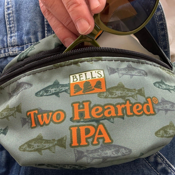 Green single pocket fanny pack with trout pattern in darker greens, with black zipper and strap. Bell's logo in orange and white as well as Two Hearted IPA printed in orange letters with green outline.  