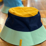 A reversible bucket hat with Oberon tag , teal brim, blue base and orange top on one side. On the other is an Eclipse tag, teal brim, dark blue base and yellow top.