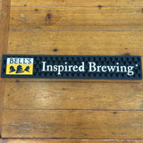Skinny rectangular black rail mat with bell texture. Yellow, black, and white logo on the left. Inspired Brewing text in white to the right.