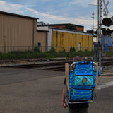 Person with a blue backpack walking near train tracks and buildings.