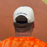 BACK VIEW OF WHITE HAT WITH BLUE BILL, AND EMBOIDERED "FOR THE LAKES" ABOVE STRAP