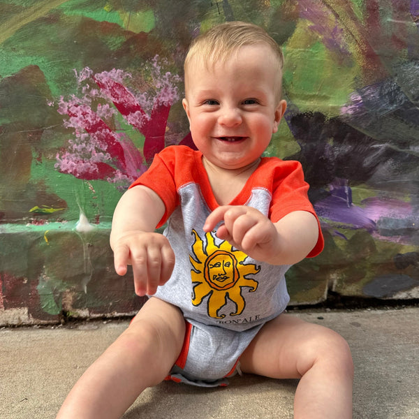 A grey baby onesie with a raglan style orange sleeve and accents.  Features a yellow Oberon sun on the front.