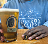 A person wearing a blue long-sleeve shirt with glow-in-the-dark 'OBERON ECLIPSE' and a starry space graphic holds a pint of beer in a clear glass printed withfull color 'OBERON ECLIPSE' logo. The person's hands are visible, along with a small OBERON ECLIPSE branded enamel pin on the table.