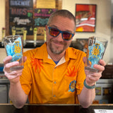 Man wearing blue Oberon sunglasses and an orange Oberon button shirt holding two pint glasses in a bar setting.