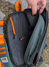 A close-up of a green and orange sling bag being opened by a hand, showing the interior compartments, zippers, and a small attached black carabiner. Part