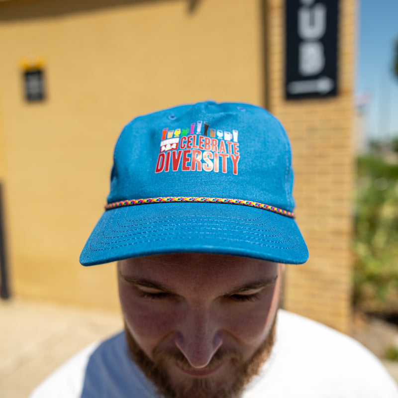Blue canvas hat with Bell's Celebrate Diversity logo in red.  Above that is a rainbow assortment of different beer style glassware, 11 styles ranging from red to gray. Also featuring rainbow rope between the bill and crown of hat