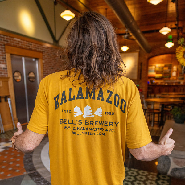 golden yellow tee shirt with Kalamazoo printed in college style on the back in charcoal black with "ESTD  1985" alongside white Bells logo bells. also includes name and address in charcoal black.