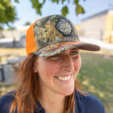 a trucker hat with orange mesh and hunter's camo front and bill featuring Deer Camp logo patch