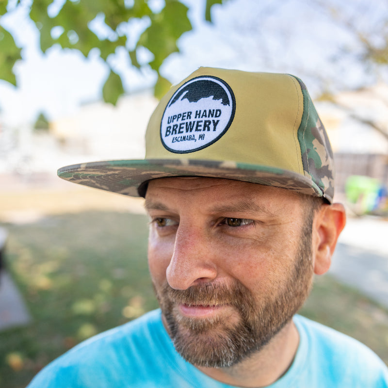 camo hat and solid beige front with black and white circular Upper Hand Brewery logo patch