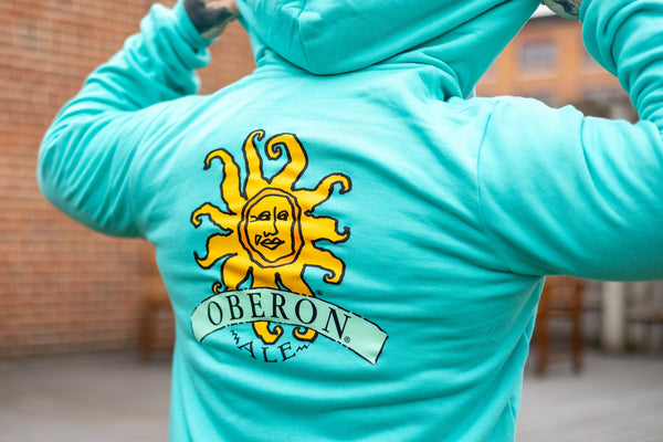 Teal hoodie, large yellow and orange Oberon sun logo on the back.