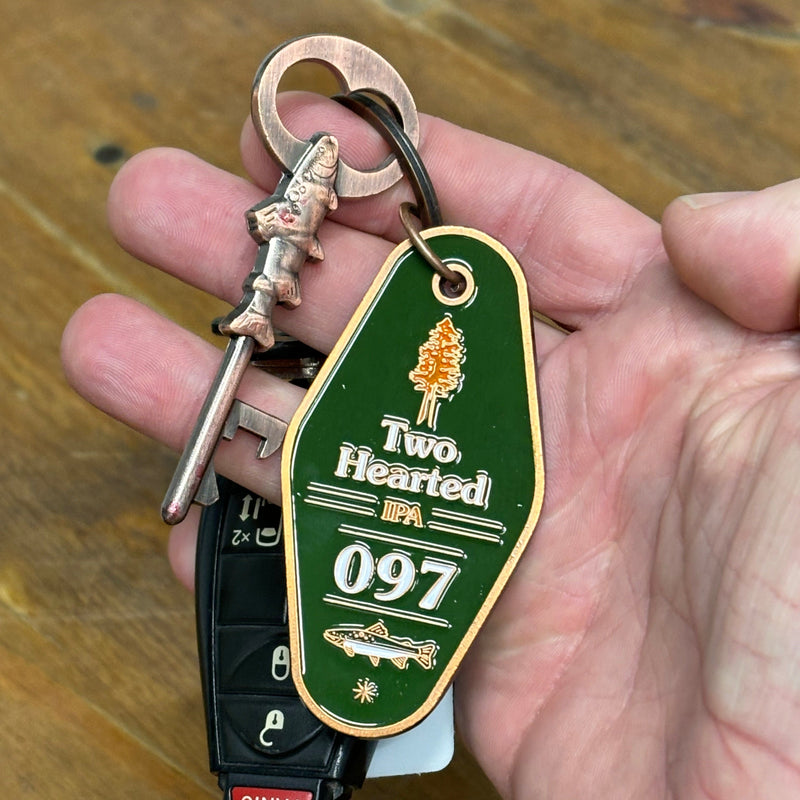 copper color keychain with green enamel paint front side featuring and Orange pine tree, Two Hearted IPA and 097 in white, with the two hearted fish below in white and green.  there is an additional old timey key with a trout at the base and a bottle opener at the tip