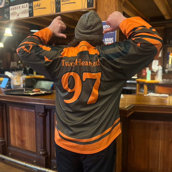 back view of Two Hearted hockey jersey, olive green base color with large orange stripes at the bottom of the body and near the end of the sleeves.  The top panel around the neck and shoulder is a green and orange camo.  Blow that is "Two Hearted" in orange, and the large number 97 in orange with a dark green border