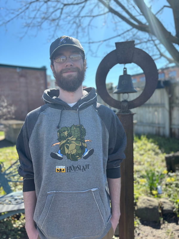 Gray hoodie with black sleeves, hood, and drawstrings. Printed on the front is the Bell's Brewery Hopslam logo underneath an illustration of 3 large hop plants slamming into a person in jeans and sneakers.