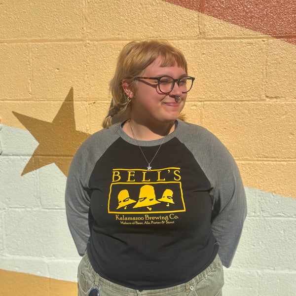 Person wearing a black and gray 3/4 sleeve baseball tee with vintage 'Bells' logo in front of a colorful wall.