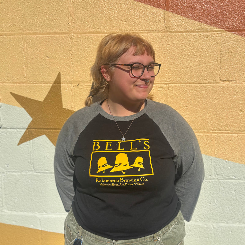 Person wearing a black and gray 3/4 sleeve baseball tee with vintage 'Bells' logo in front of a colorful wall.