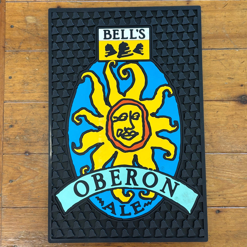 Black bar serve mat with bell texture. Features the Oberon Ale logo in various blues, yellow, and orange. Bell's logo in yellow, black and white centered at the top.