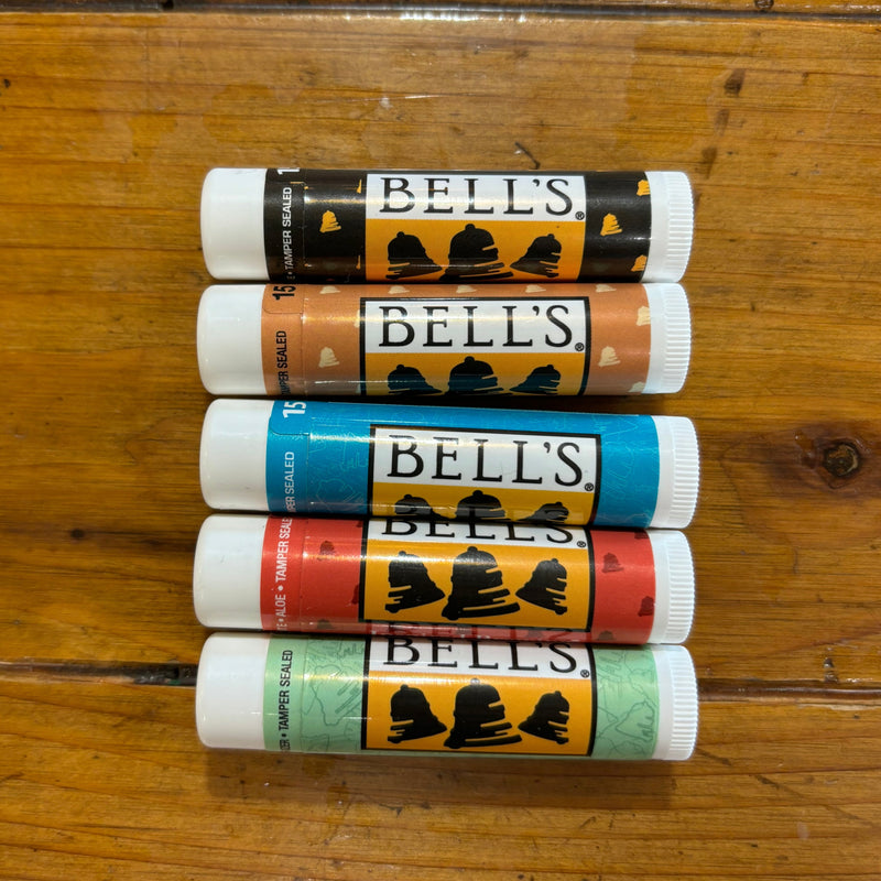 Variety pack of lip balm tubes. From left to right, light blue, black, red, yellow, tan, light green.. All feature the black, white, and yellow Bell's logo. All have tamper teals.