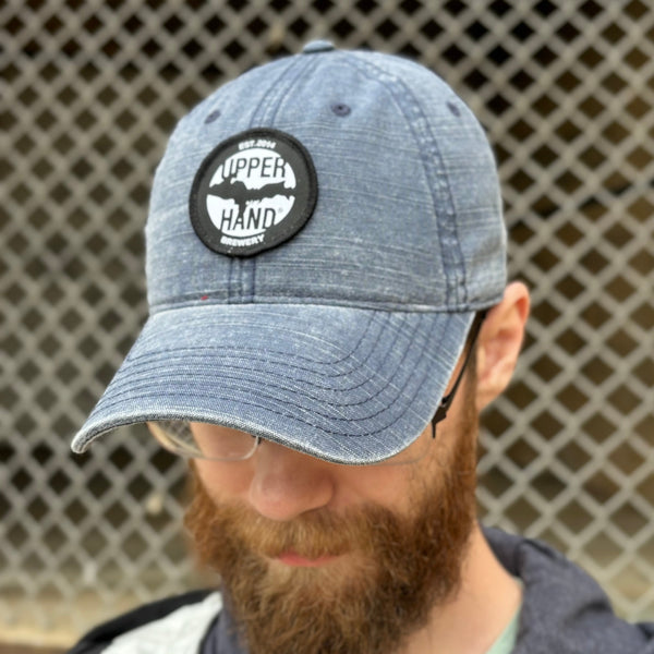 A washed navy cotton twill dad hat style baseball cap with a curved brim and adjustable fabric strap closure. Sewn onto the front panel is an embroidered patch featuring the Upper Hand Brewery logo.  