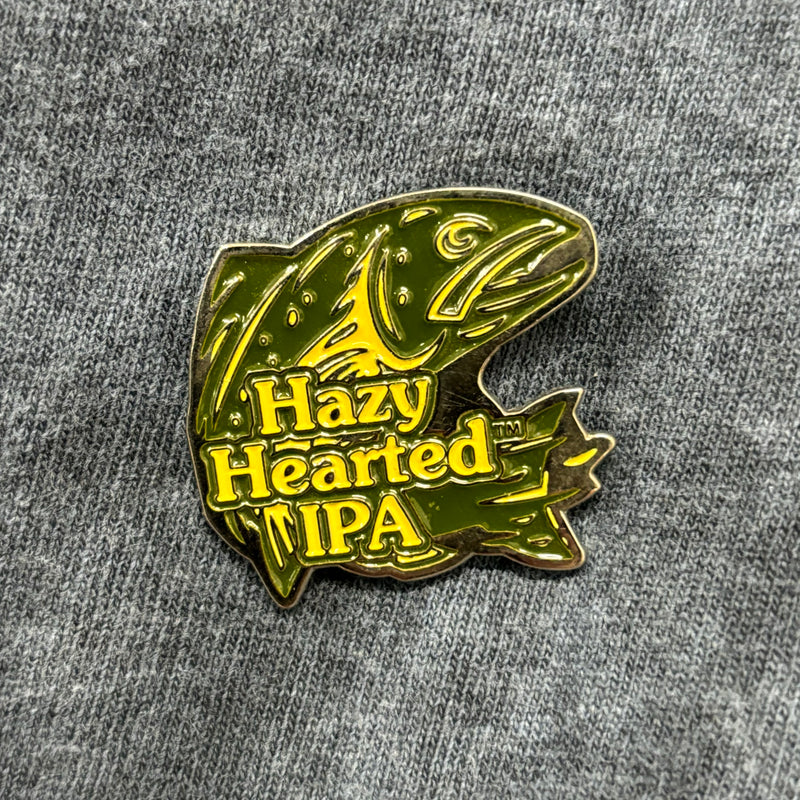 Enamel pin of Hazy Hearted logo, featuring a green trout with yellow accents, and the words Hazy Hearted IPA in yellow.