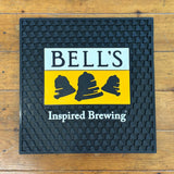 Black server mat with bell texture. Features a yellow, black, and white Bell's logo in the center with "Inspired Brewing" in white centered below the logo.