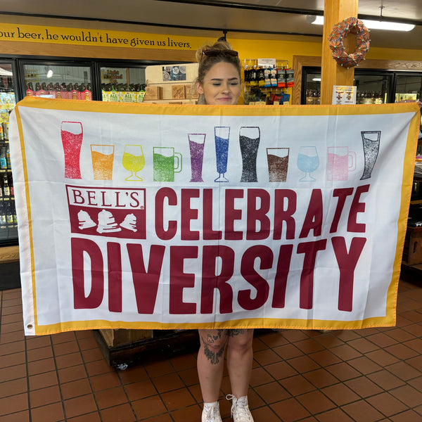 White with a thick yellow border, rectangle tin sign with eleven beer glasses filled with liquids of rainbow colors, including (Left to Right), red, orange, yellow, green, purple, blue, black, brown, light blue, pink, and gray. "Celebrate Diversity" and Bell's logo below in maroon.