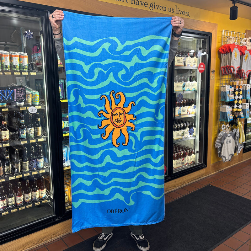 beach towel with 2 color wave pattern, blue and teal.  Full color Oberon sun in the center, and Oberon spelled out at the bottom in black