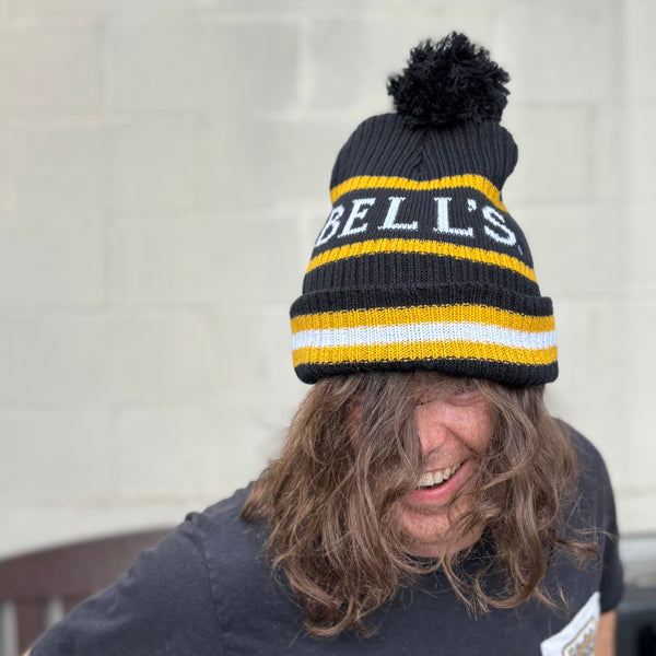Person wearing a black and yellow striped beanie with 'BELL'S' branding in white, with black pom.