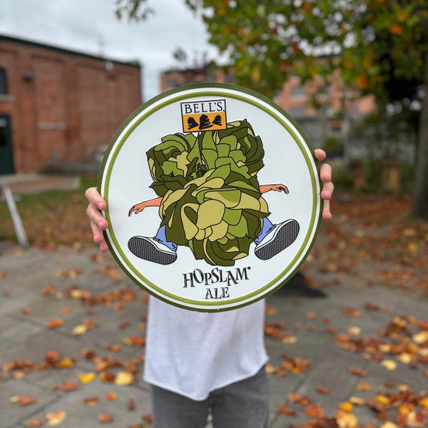 Round sign with green borders, the Bell's Brewery logo on top, illustration of 3 hop plants slamming into a person with their arms and legs sticking out, and the Hopslam Ale logo underneath.