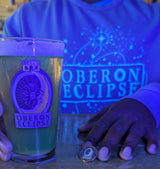 A person wearing a blue long-sleeve shirt with glow-in-the-dark 'OBERON ECLIPSE' and a starry space graphic holds a pint of beer in a clear glass printed withfull color 'OBERON ECLIPSE' logo. The person's hands are visible, along with a small OBERON ECLIPSE branded enamel pin on the table.