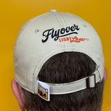 Off white baseball hat with Flyover Light Lager text branding in blue and orange with buckle strap and pinch tag with Bell's logo
