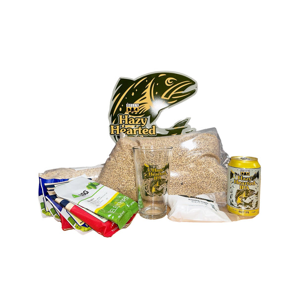 Hazy Hearted IPA fish-shaped tin sign in green and yellow. Kit ingredients displayed in front, including hops, pint glass, priming sugar, grain, and a can of Hazy Hearted IPA