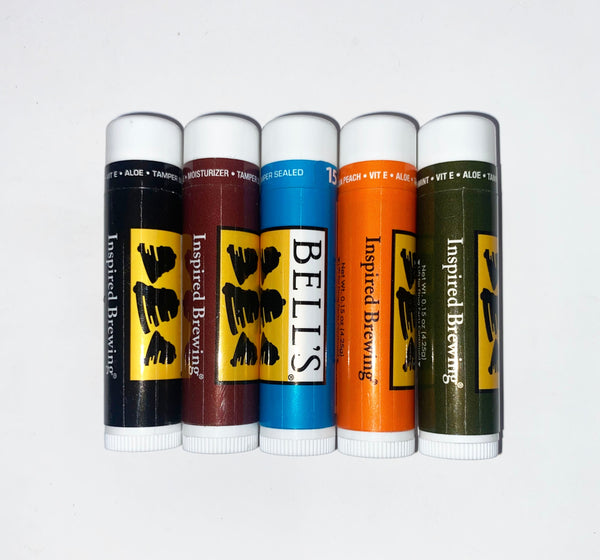 Variety pack of lip balm tubes. From left to right, Black wrapper, Brown wrapper, light blue wrapper, orange wrapper, green wrapper. All feature language "Inspired Brewing" and the black, white, and yellow Bell's logo. All have tamper teals.