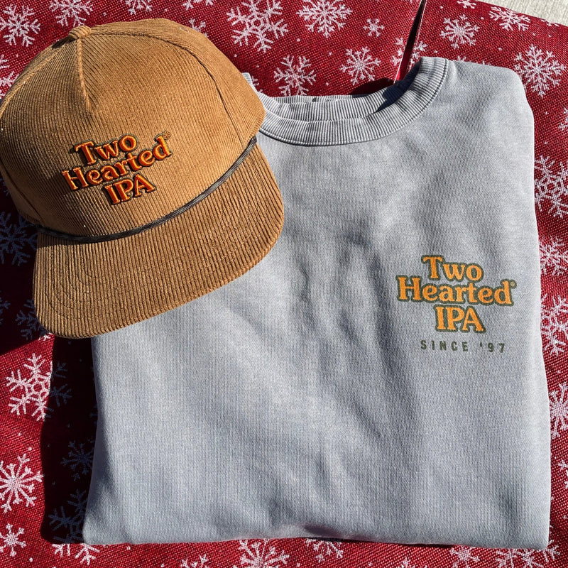 Gray Two Hearted crewneck sweater, with Two Hearted IPA printed in orange letters with green outline, on the pocket. beneath is "Since '97" in smaller green letters. Image also features a brown corduroy hat with "Two Hearted IPA" embroidered in orange with green outline