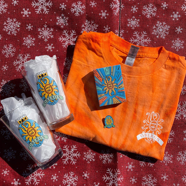 Orange tie dye t shirt with small white outline Oberon logo on front pocket, and again large on the back.  2 glass 16oz pints with full color Oberon logo, one deck of playing cards with Oberon Sunburst logo package, and one full color Oberon logo enamel pin