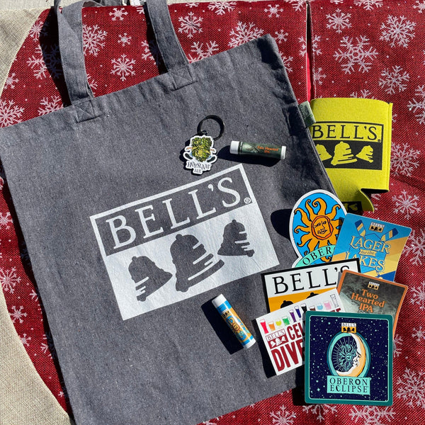 gray tote bag with Bell's logo in white, with an assortment of Bell's brand stickers and Hopslam keychain, Two Hearted LIp balm, Oberon lip balm, and logo printed can sleeve