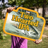 Rectangular-like Two Hearted IPA tin sign featuring the Two Hearted fish artwork and Two Hearted IPA text. Yellow, black and white Bell's logo top center. Bottom text in pale yellow: American India Pale Ale - Bell's Brewery, Inc. - Comstock, MI. Has an orange border around sign.