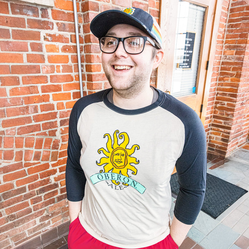 oat colored baseball tee shirt with black sleeves and neck, featuring large full color Oberon logo on front