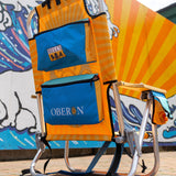 beach chair with light orange and dark orange sunburst from center. Center back features Oberon Ale logo. Teal head rest pillow with Bell's logo at the top of the chair.  Rearview of chair shows 2 large blue zip up storage pouches on the back of chair, top one with Bell's logo, and bottom with Oberon spelled across.