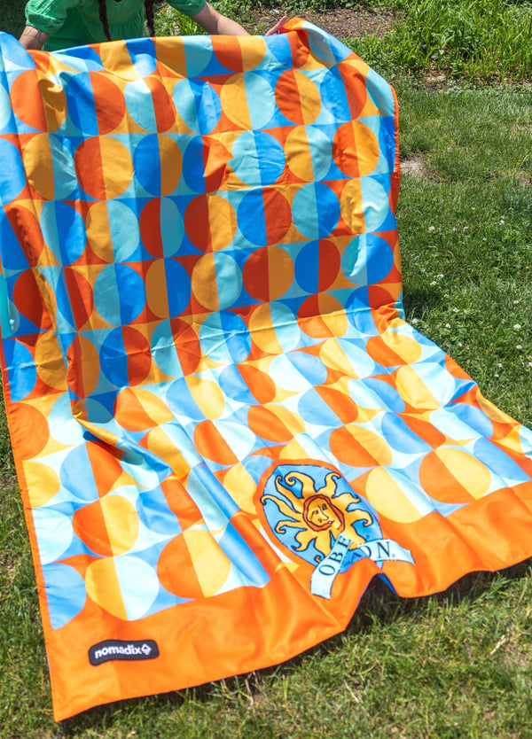 Orange framed circular patterns with alternating colors, light and dark blues as well as gold and orange.  Underside is full blue.  Oberon logo large center placement at the bottom of blanket.