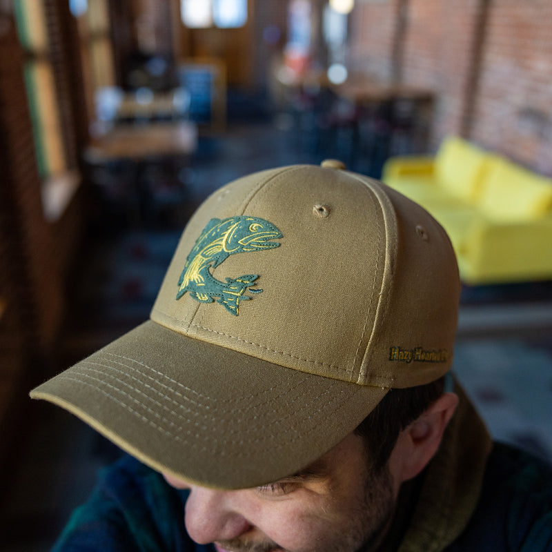 A khaki baseball hat with a green and yellow fish patch on on the front and a "Hazy Hearted" patch on the side. 