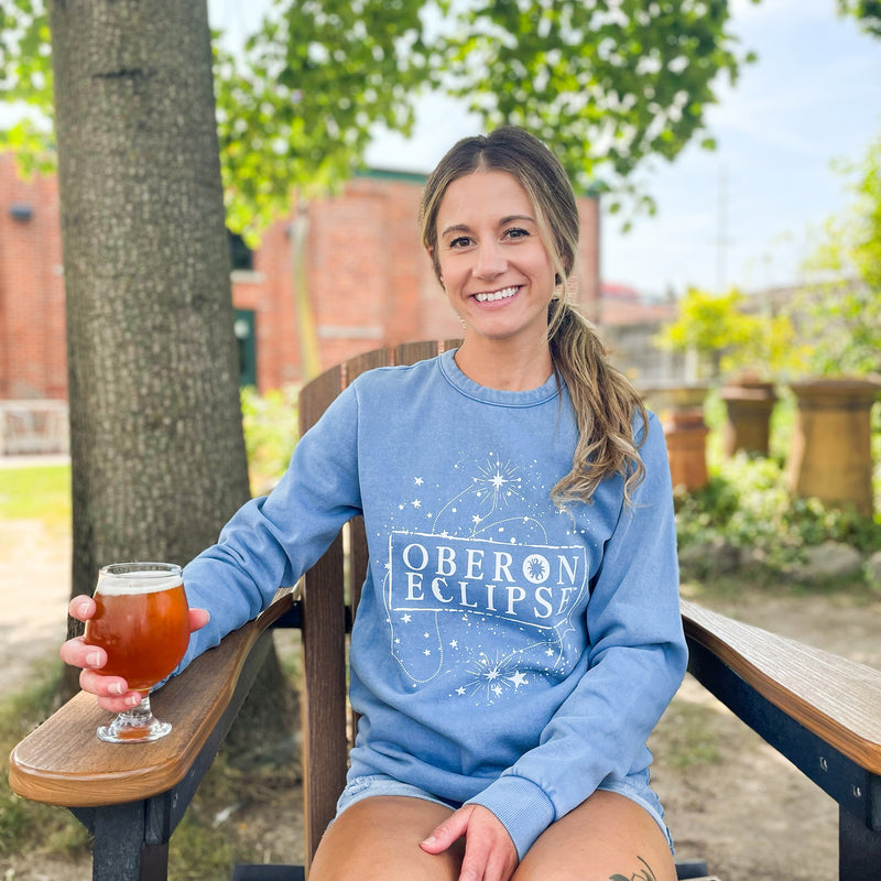 light denim blue crewneck sweater with glow painted Oberon Eclipse logo with night sky motif in glow paint on front