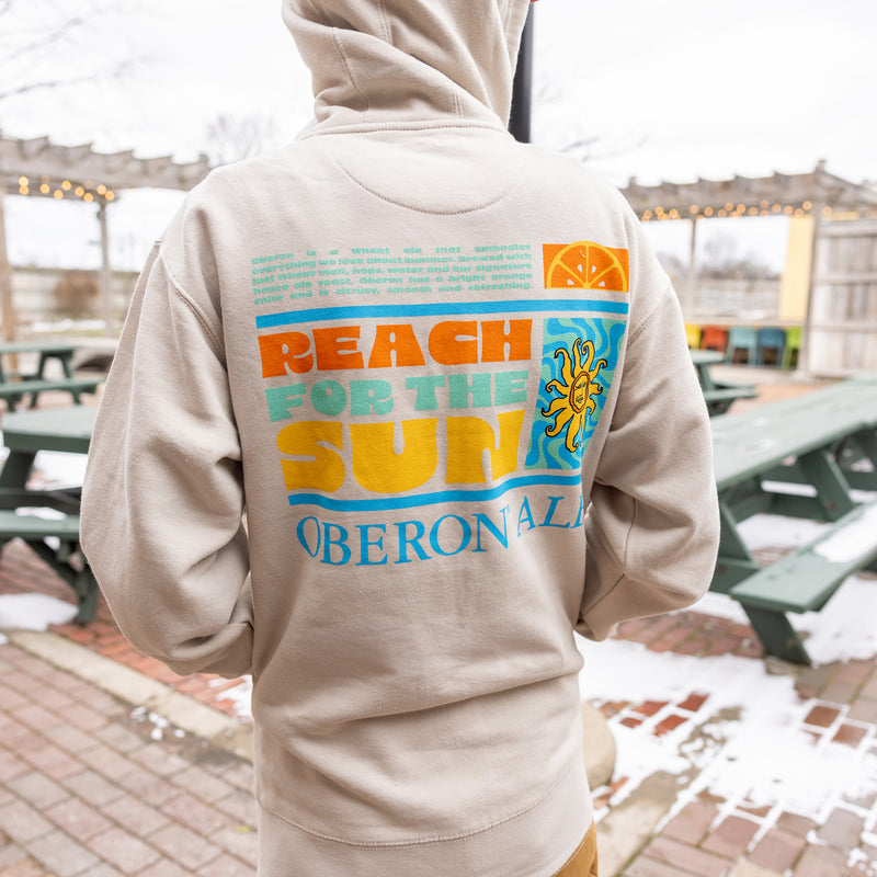 Back view of bone white zip up hoodie with "Reach for the sun" spelled out in Oberon colors next to Oberon logo on wave graphic background.  There is a beer description above main graphic in see green with an orange slice next to it.  Below the main graphic is Oberon le spelled out in blue