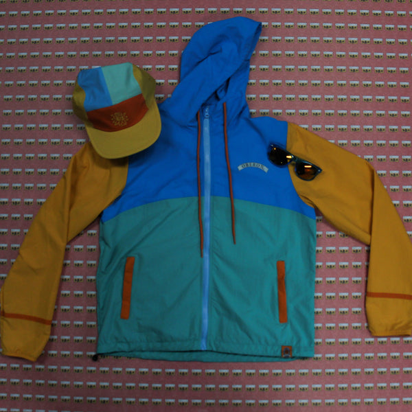 Blue, sea green, yellow and orange color block windbreaker jacket with a color block hat and blue sunglasses 