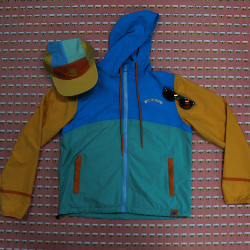 Blue, sea green, yellow and orange color block windbreaker jacket with a color block hat and blue sunglasses 