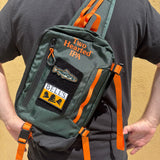 A dark green sling backpack with bright orange straps and patches. The backpack features 'Two Hearted IPA' embroidered on the front, a fish patch, and a 'BELLS' patch.