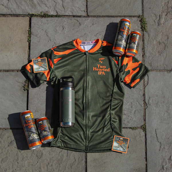Green and orange Bike Jersey, camo sleeves and with 'Two Hearted IPA' branding, surrounded by beer cans and a Two Hearted Yeti forest Green water bottle on a stone surface.